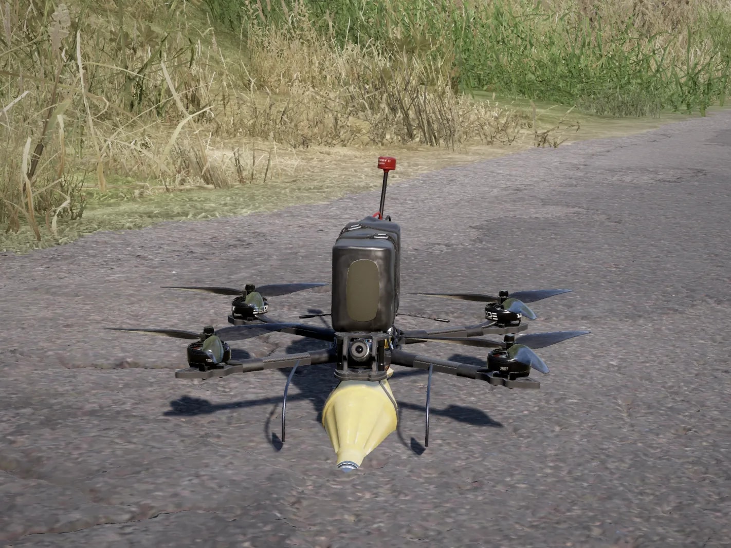 Drone FPV kamikaze visto de frente, com bateria em cima e ogiva amarela de RPG presa embaixo, pousado em uma estrada