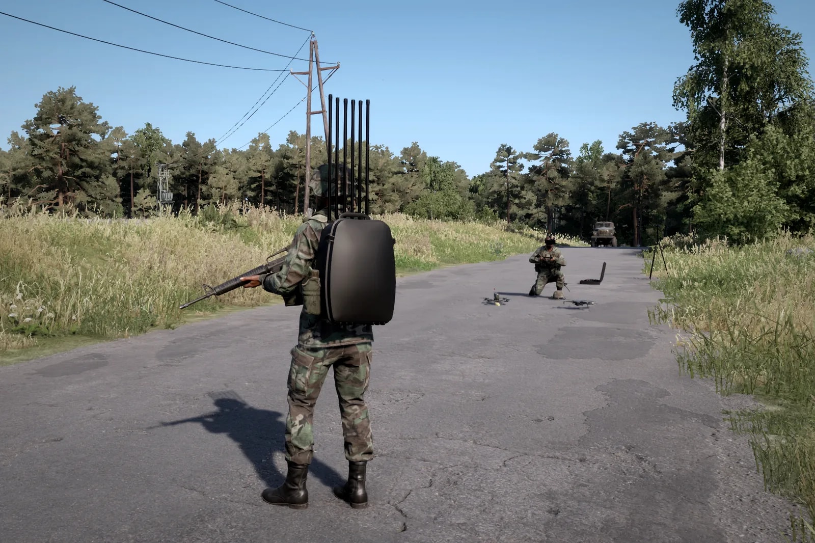 Soldado de costas com a mochila jammer e suas antenas verticais, a poucos metros de um operador de drone ajoelhado na estrada