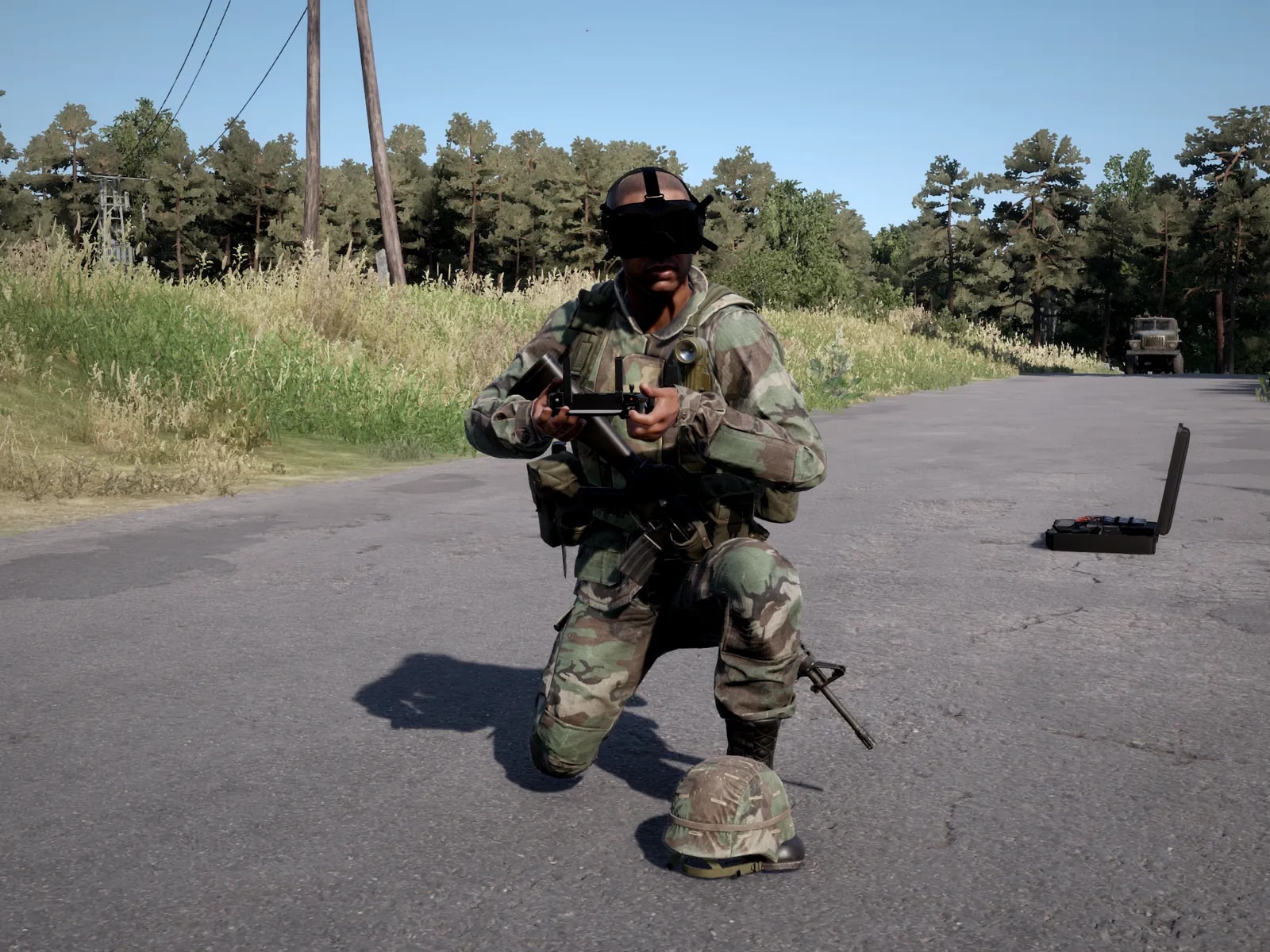 Soldado ajoelhado na estrada com os óculos FPV no rosto e o controle do drone nas mãos; o capacete fica no chão ao lado