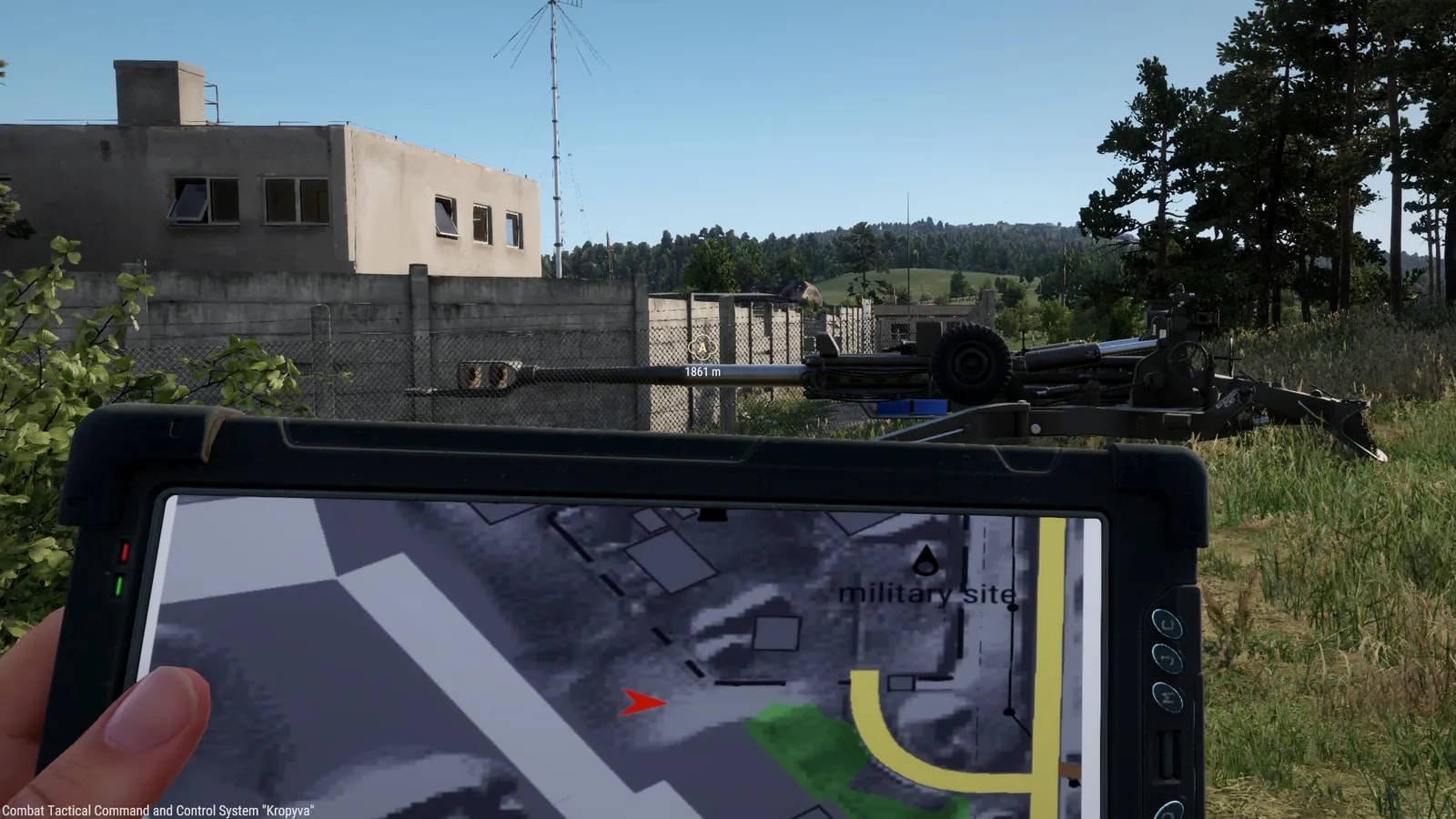 Visão em primeira pessoa de um soldado segurando o tablet Kropyva na mão esquerda, com o mapa da área na tela e uma seta vermelha no centro; atrás, um obus M777A2 sobre a grama ao lado de um muro de concreto